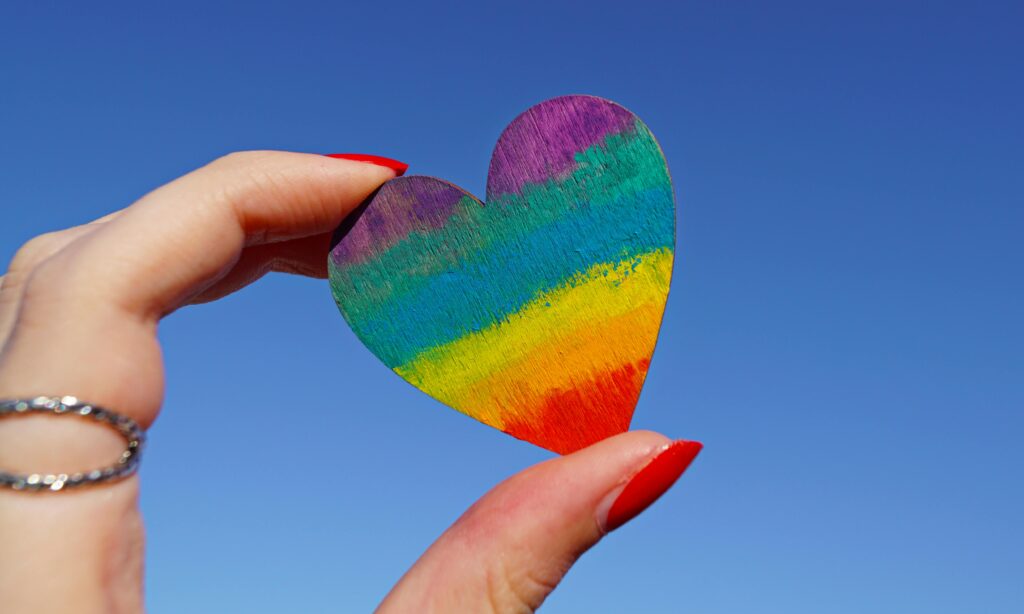 A hand holding a rainbow-colored heart against a clear blue sky, symbolizing love and pride.